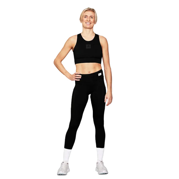 En kvinde står på en hvid baggrund iført en sort sports-BH, Saysky Motion+ lange tights, hvide sokker og hvide sportssko. Hun har den ene hånd på hoften, mens hun smiler.