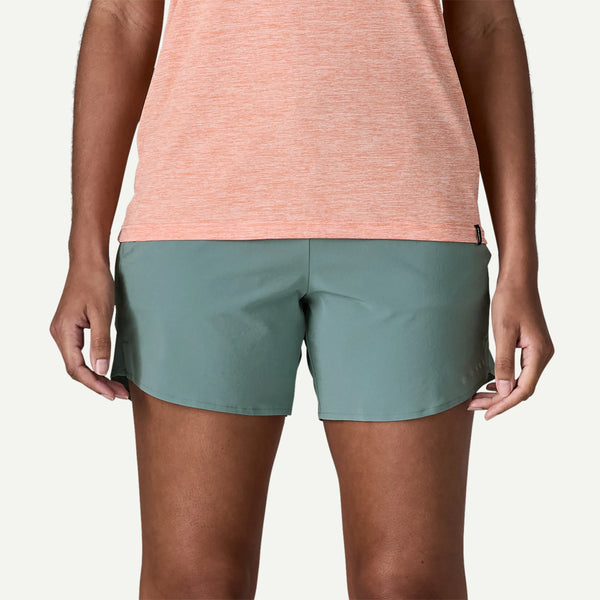 En person iført grønne Patagonia Multi Trails Shorts 5½in, Dame in Blue Sage og en ferskenfarvet kortærmet skjorte står med ansigtet fremad og afslappede arme; billedet er beskåret fra midt på torsoen til midt på låret.