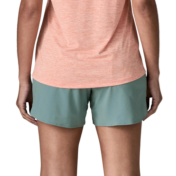 En person står med ryggen til kameraet iført en ferskenfarvet kortærmet skjorte og blå Patagonia Multi Trails Shorts 5½in, Dame. Kun overkroppen og benene er synlige.
