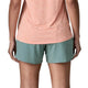 En person står med ryggen til kameraet iført en ferskenfarvet kortærmet skjorte og blå Patagonia Multi Trails Shorts 5½in, Dame. Kun overkroppen og benene er synlige.