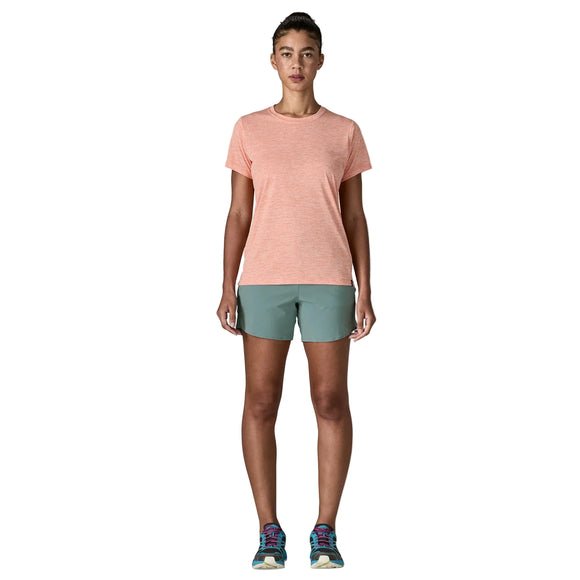En person står med front mod en lys orange kortærmet skjorte, Patagonia Multi Trails Shorts 5½in til kvinder i Blue Sage og løbesko på en almindelig hvid baggrund.