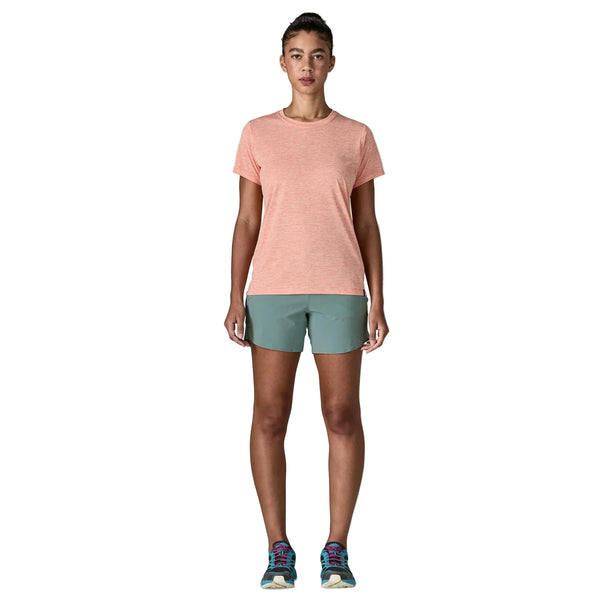 En person står med front mod en lys orange kortærmet skjorte, Patagonia Multi Trails Shorts 5½in til kvinder i Blue Sage og løbesko på en almindelig hvid baggrund.