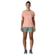 En person står med front mod en lys orange kortærmet skjorte, Patagonia Multi Trails Shorts 5½in til kvinder i Blue Sage og løbesko på en almindelig hvid baggrund.