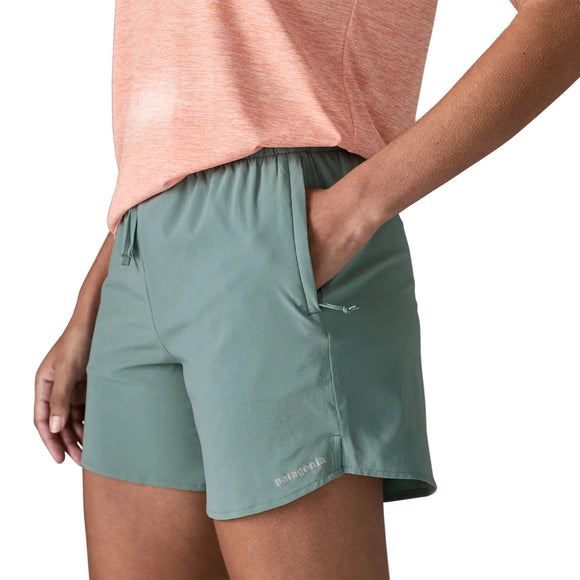 En person er iført blå salvie Patagonia Multi Trails Shorts 5½in til kvinder og en lys orange lyngfarvet T-shirt, mens han står med den ene hånd i lommen. Shortsene har elastik i taljen, snøre og et lille Patagonia-logo på venstre ben.