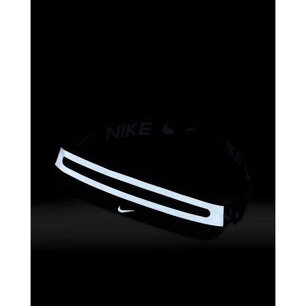 Nike Slim Waistpack 4.0 Unisex i sort/sort/sølv har en vandret refleksstribe og et lille hvidt Nike-logo på forsiden. Ideel til at bære de vigtigste ting, mens du løber. Vist mod en mørk baggrund.