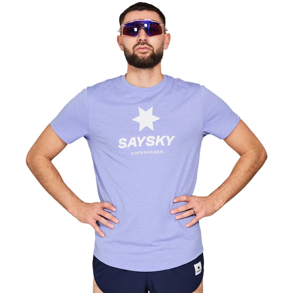 En skægget mand bærer mørke solbriller og Saysky Logo Combat T-shirt, Herre, Purple Melange med marineblå shorts. Den fugttransporterende skjorte holder ham kølig, mens han står med hænderne på hofterne mod en almindelig lysegrå baggrund.