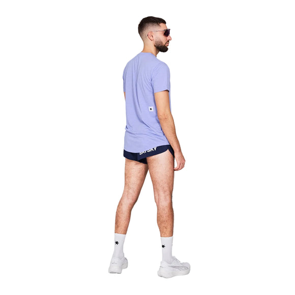 En mand med kort hår og skæg står med ryggen til i et almindeligt studie iført Saysky Logo Combat T-shirt, Herre, Purple Melange, marineblå trusser, hvide sokker, hvide sneakers og solbriller.