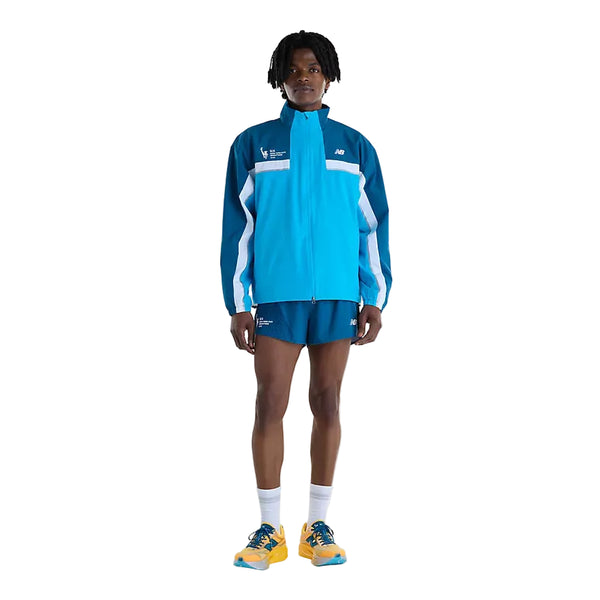 En person iført en blå og turkis New Balance NYC Marathon-jakke, Herre, Altitude-træningsjakke med lynlås og matchende løbeshorts, samt hvide sokker og gule sportssko.