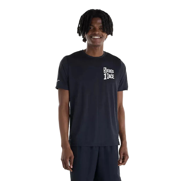 En person iført en sort New Balance NYC Marathon RC Essential Graphic T-shirt (herre) og sorte shorts står foran en hvid baggrund. T-shirten, der er udstyret med NB ICEx-teknologi, har hvid tekst på venstre bryst, hvor der står »5 BOROS 1 RACE«. Personen smiler og har kort, krøllet hår.