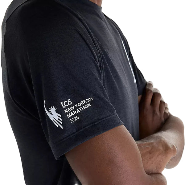 En person med armene over kors iført New Balance NYC Marathon RC Essential Graphic T-shirt til mænd i sort, med teksten »tcs NEW YORK CITY MARATHON 2026« og et stiliseret logo af Frihedsgudindens fakkel trykt på ærmet. Denne T-shirt er fremstillet med avanceret NB ICEx-teknologi, der sikrer åndbarhed og kølig komfort under hver eneste løbetur.