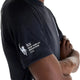 En person med armene over kors iført New Balance NYC Marathon RC Essential Graphic T-shirt til mænd i sort, med teksten »tcs NEW YORK CITY MARATHON 2026« og et stiliseret logo af Frihedsgudindens fakkel trykt på ærmet. Denne T-shirt er fremstillet med avanceret NB ICEx-teknologi, der sikrer åndbarhed og kølig komfort under hver eneste løbetur.