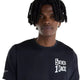 En person med mellem-mørk hud og korte dreadlocks har en New Balance NYC Marathon RC Essential Graphic T-shirt til mænd i sort på, med påskriften »5 BOROS 1 RACE«. Billedet er taget fra brystet og opefter, hvor personen står med ansigtet vendt mod kameraet foran en ensfarvet hvid baggrund.