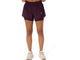 En person står med ansigtet fremad og har Asics Nagino Run 2-N-1-shorts til kvinder i farven Dark Aubergine på, med et lille logo på venstre ben, en hvid skjorte, der er stoppet ned i bukserne, og beige sportssko. Billedet er beskåret ved taljen og anklerne.