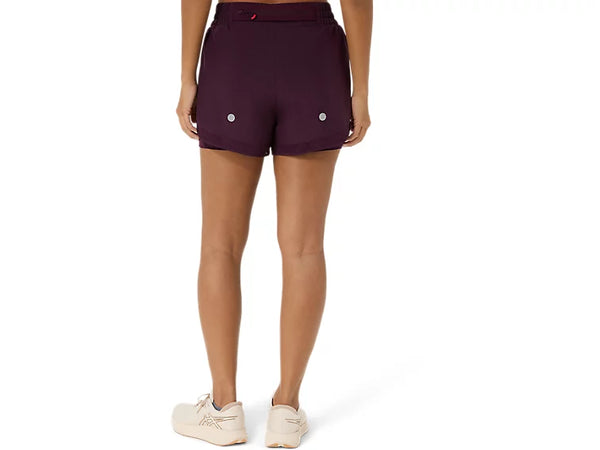 En person står med ryggen til og har Asics Nagino Run 2-N-1-shorts til kvinder i mørk auberginefarve på, med to reflekterende prikker på bagsiden, samt hvide sneakers. Baggrunden er ensfarvet hvid.