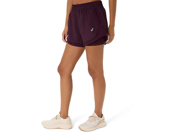 En person står i en neutral stilling iført Asics Nagino Run 2-N-1-shorts til kvinder i farven Dark Aubergine, der er forsynet med et lille Asics-logo, sammen med en matchende top og lyse sportssko på en ensfarvet hvid baggrund. Nagino Run 2-N-1-shortsene fra Asics er designet til at give komfort og stil til hver eneste løbetur.