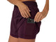 En kvinde iført mørke, auberginefarvede Asics Nagino Run 2-N-1-shorts lægger en smartphone i en sidelomme. Disse vandafvisende løbeshorts til kvinder har en elastisk linning og vises på en ensfarvet hvid baggrund.