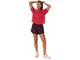 En person står med armene løftet og er iført en løs rød T-shirt, Asics Nagino Run 2-N-1-shorts til kvinder i farven Dark Aubergine samt lyse sneakers. Baggrunden er helt hvid.