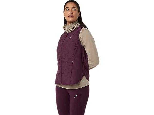 En person står med ansigtet vendt mod venstre og er iført en beige langærmet bluse, matchende rødbrune leggings og ASICS Nagino Run Padded Gilet til kvinder i farven Dark Aubergine. Løbevesten til kvinder fra ASICS har et atletisk design og fremstår på en ensfarvet hvid baggrund.