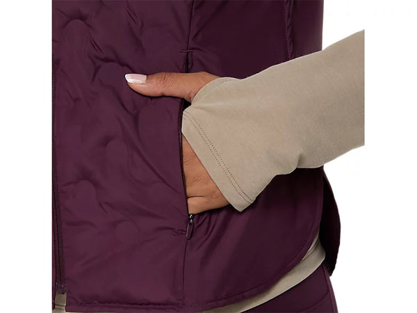 En person iført en beige langærmet bluse stikker hånden ned i lommen på en mørk auberginefarvet Nagino Run Padded Gilet (Dame) fra Asics. Kun overkroppen og en del af armen er synlige på en ensfarvet baggrund.
