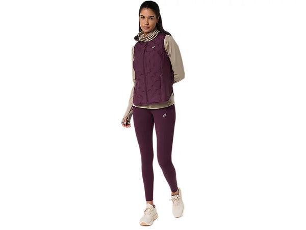 En person står og kigger fremad. Hun har en beige langærmet bluse på, nemlig ASICS Nagino Run Padded Gilet til kvinder i farven Dark Aubergine, lilla leggings og hvide sneakers. Baggrunden er ensfarvet hvid.