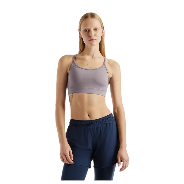 En person med langt, glat hår har en New Balance Nb Essential Yoga Bra, Dame, i farven Off-Line Grey på og marineblå sportshorts og står foran en hvid baggrund.