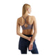 Der ses en person med langt blondt hår set bagfra, iført en New Balance Nb Essential Yoga Bra, Dame, i farven Off-Line Grey samt marineblå sportshorts, mens vedkommende med højre hånd holder sit venstre håndled bag ryggen på en hvid baggrund.