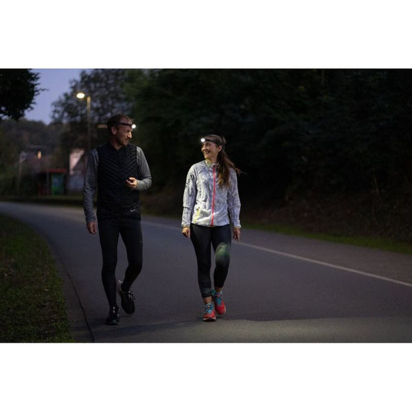 En mand og en kvinde i sportstøj, som begge bruger Led Lenser Neo 3 400 Lumens Unisex Black pandelampe, går og smiler sammen under deres natlige løbetur på en vej med træer og gadebelysning.