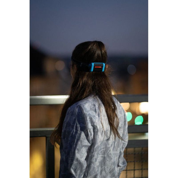 En person med langt brunt hår står på en balkon i skumringen, iført en mønstret jakke og en Led Lenser Neo 3 400 Lumens, Unisex, Black pandelampe, klar til et aftenløb.