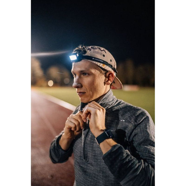 Iført en Led Lenser Neo 5R 600 Lumens pandelampe og en kasket lyner en person en grå jakke op på en udendørs løbebane om natten, og det sorte unisex-lys oplyser den dunkle sti til et sent aftenløb.