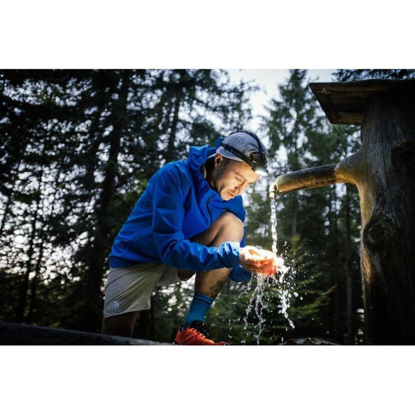 En person iført blå jakke og Led Lenser Neo 9R 1200 Lumens Unisex-pandelampe i sort sidder på hug ved en skovbrønd og fanger vand i hænderne, mens sollyset filtreres gennem træerne - ideelt til trailløbseventyr.