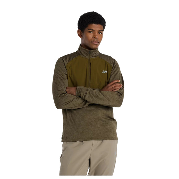 En person står med armene over kors iført en grøn New Balance Athletics Heat Grid 1/2 Zip for men in Woodland Heather og beige bukser på en hvid baggrund.