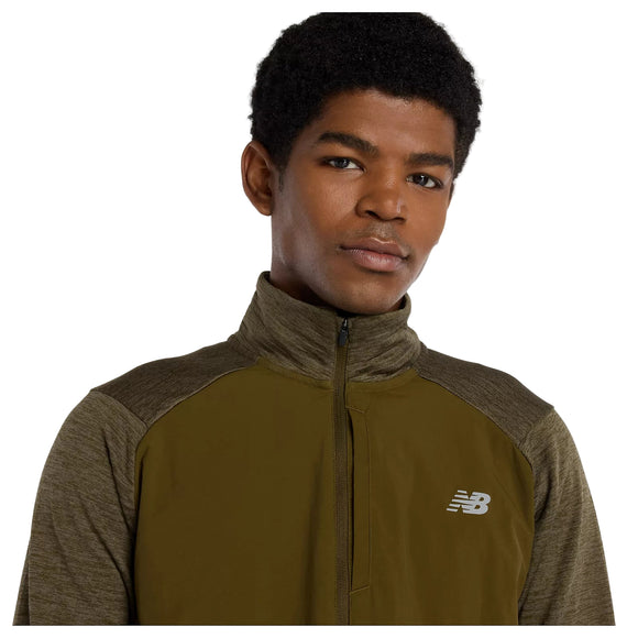 En person med kort, krøllet hår bærer en New Balance Athletics Heat Grid 1/2 Zip, Herre, Woodland Heather, og kigger direkte ind i kameraet mod en almindelig hvid baggrund.