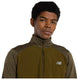 En person med kort, krøllet hår bærer en New Balance Athletics Heat Grid 1/2 Zip, Herre, Woodland Heather, og kigger direkte ind i kameraet mod en almindelig hvid baggrund.