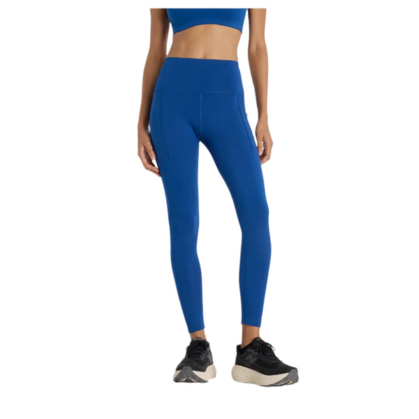En kvinde bærer New Balance Sleeck Pocket High Rise Tights, Dame, Galaxy Blue, med en matchende blå sports-bh og sort-hvide sneakers på en hvid baggrund. De fugttransporterende tights er ideelle til aktiv træning. Hendes hoved er ikke synligt.