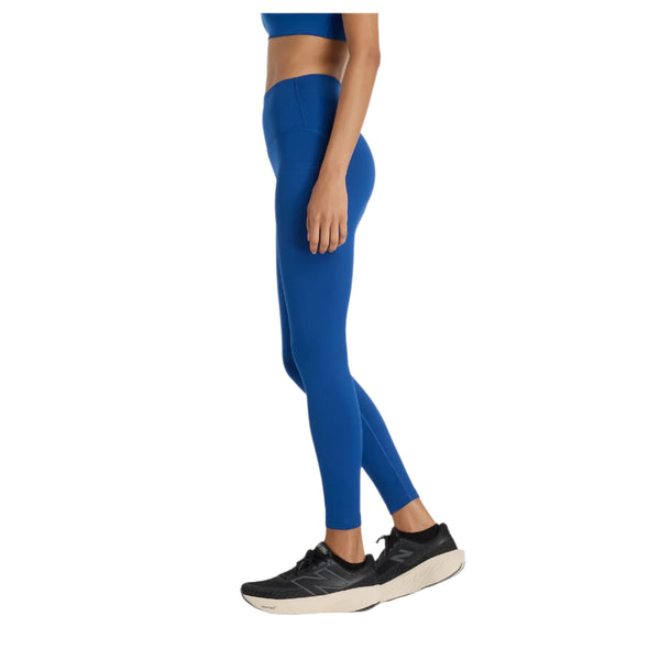 En person, der er synlig fra skuldrene og ned, står sidelæns i en blå sports-bh og New Balance Sleeck Pocket High Rise Tights, Dame, Galaxy Blue, sammen med sorte sportssko på en almindelig hvid baggrund.