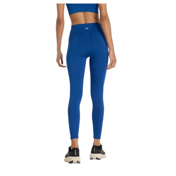 En person står med front mod kameraet iført en blå sports-bh, New Balance Sleeck Pocket High Rise Tights til kvinder i Galaxy Blue og sorte og hvide sportssko på en almindelig hvid baggrund.