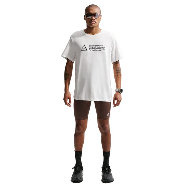 En person står og kigger fremad iført solbriller, en Nike ACG Df Tee Std Trail Logo til mænd i Summit White med sort trekantlogo og tekst, brune, tætsiddende shorts, sorte sokker og sorte sneakers på en almindelig hvid baggrund.