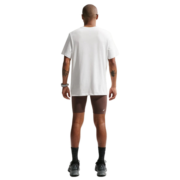 En person står med front mod kameraet iført Nike ACG Df Tee Std Trail Logo, Herre, Summit White, brune, tætsiddende shorts, sorte sokker og sorte sportssko på en hvid baggrund.