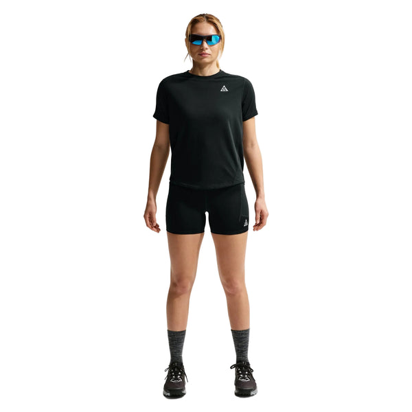 En kvinde kigger fremad i Nike ACG High-Waisted 4" Running Shorts, Dame, Black/Summit White, sammen med en sort t-shirt, grå sokker, sorte sportssko og blå solbriller. Baggrunden er helt hvid.
