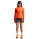 En person står med ansigtet vendt fremad iført en orange langærmet trøje, Nike ACG High-Waisted 4″ løbeshorts til kvinder i farverne Ironstone/Cave Stone/Summit White, hvide sokker, orange sneakers og solbriller. Baggrunden er helt hvid.