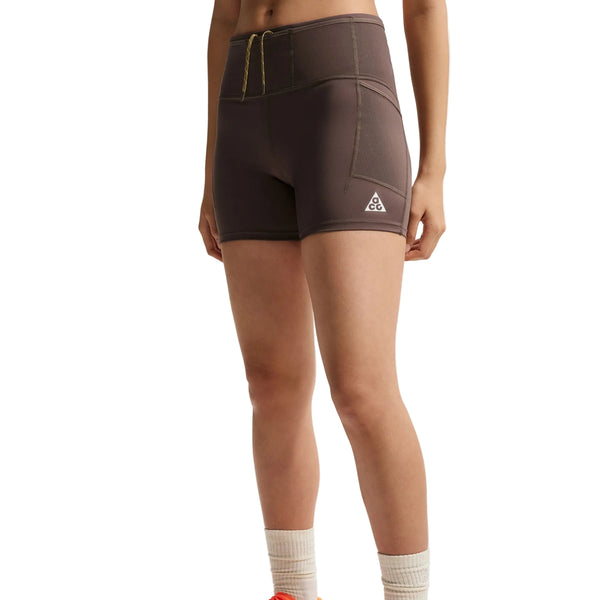 En person står foran en hvid baggrund iført Nike ACG High-Waisted 4″ løbeshorts til kvinder i farverne Ironstone/Cave Stone/Summit White, med et lille hvidt logo på venstre ben. Personens ansigt kan ikke ses.