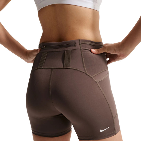 En person har Nike ACG High-Waisted 4″ løbeshorts til kvinder i farverne Ironstone/Cave Stone/Summit White på, med synlige sømme og en lynlåslomme bagpå, set fra taljen og ned og bagfra. Personen har desuden en hvid sports-BH på og har begge hænder hvilende på hofterne.
