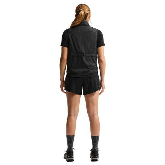 En person med langt tilbagestrøget hår, set bagfra, er iført en sort kortærmet skjorte, sort vest, Nike ACG Mid-Rise Brief-Lined Running Shorts (Dame, Black/Black/Summit White), sorte sokker og sorte sportssko på en hvid baggrund.