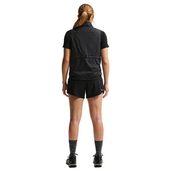 En person med langt tilbagestrøget hår, set bagfra, er iført en sort kortærmet skjorte, sort vest, Nike ACG Mid-Rise Brief-Lined Running Shorts (Dame, Black/Black/Summit White), sorte sokker og sorte sportssko på en hvid baggrund.
