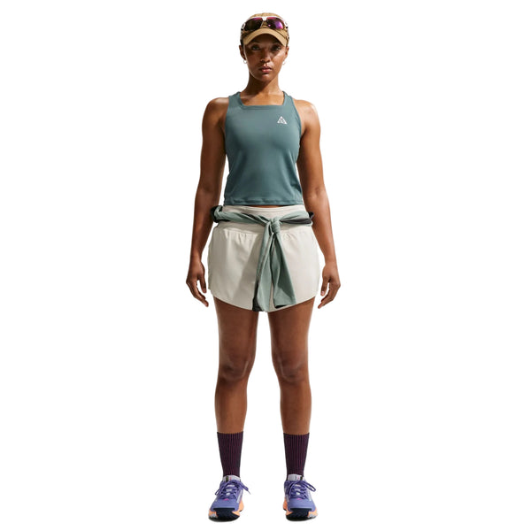 En person står med ansigtet vendt fremad og er iført en grøn tanktop og Nike ACG Mid-Rise Brief-Lined løbeshorts til kvinder i farvekombinationen Sea Glass/Sea Glass/Summit White, med en langærmet trøje bundet om taljen, mørke strømper, løbesko, en kasket og solbriller. Baggrunden er ensfarvet hvid.