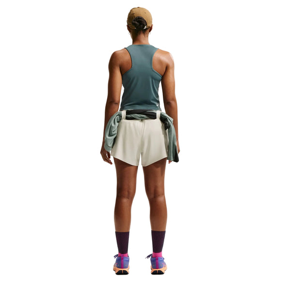 En person står med ryggen til kameraet og er iført en beige kasket, en grøn tanktop, Nike ACG Mid-Rise løbeshorts med indvendig underbuks til kvinder i farverne Sea Glass/Sea Glass/Summit White, en jakke bundet om taljen, lilla sokker samt lyserøde og orange sportssko, foran en hvid baggrund.
