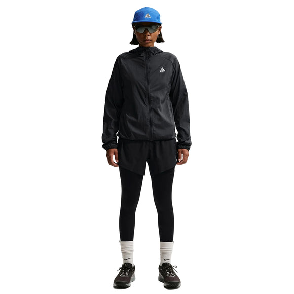 En person iført Nike ACG Repel Running Jacket til kvinder i Black/White/Summit White, sorte shorts over sorte leggings, hvide strømper, sorte sneakers, en blå kasket og sorte solbriller står på en hvid baggrund.