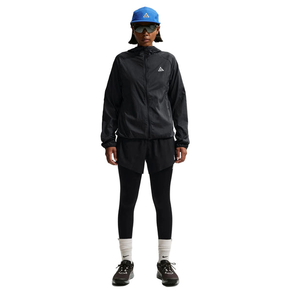 En person iført Nike ACG Repel Running Jacket til kvinder i Black/White/Summit White, sorte shorts over sorte leggings, hvide strømper, sorte sneakers, en blå kasket og sorte solbriller står på en hvid baggrund.