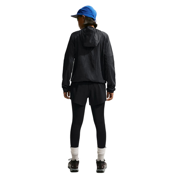 En person kigger væk, iført blå kasket, Nike ACG Repel Running Jacket til kvinder i Black/White/Summit White, sorte shorts over leggings, hvide sokker og sorte sko på en hvid baggrund.