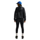 En person kigger væk, iført blå kasket, Nike ACG Repel Running Jacket til kvinder i Black/White/Summit White, sorte shorts over leggings, hvide sokker og sorte sko på en hvid baggrund.
